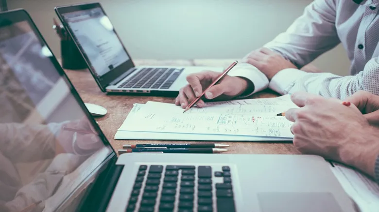 pessoa segurando lápis em uma mesa com notebooks e ao lado de uma pessoa, gestão de provas