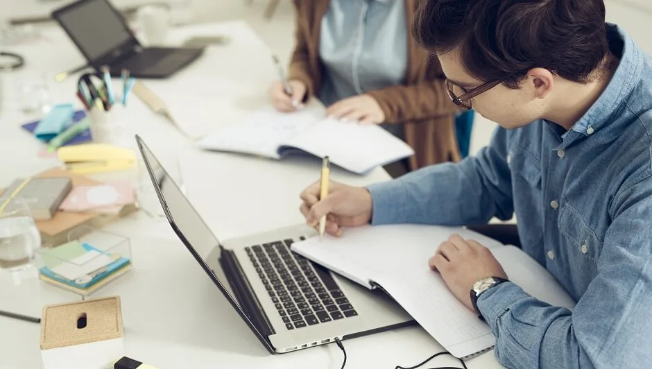 homem sentado na mesa com notebook e escrevendo no caderno